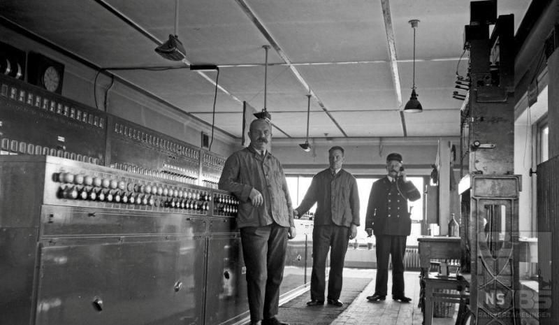 De stationschef aan de telefoon - geënsceneerd voor de foto of niet, het is niet meer te achterhalen - met twee van zijn medewerkers in post 1 te Amersfoort. We zien links het tweerijig stelknoptoestel, in die tijd zeer modern. Wat verder opvalt is de sobere inrichting van deze werkruimte. De datum is 10 augustus 1935.