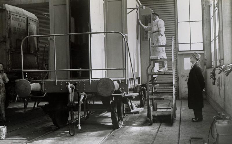 Een conducteurswagen wordt gerepareerd. Hierin reed een conducteur mee in een goederentrein; vanuit een soort erkertje kon hij langs de trein kijken. In de tijd dat deze foto werd gemaakt, was er al discussie over het nut van deze wagens. Ze zijn niet lang daarna buiten gebruik geraakt.In deze dagen was het nog gebruikelijk dat een chef een hoed droeg, terwijl handarbeiders een pet of geen hoofddeksel droegen.