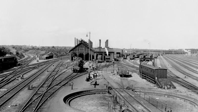 De locomotievenloods aan de westkant van Amersfoort. Voor de loods bevindt zich de draaischijf waarop men zonodig de locomotieven kon keren.Links gaat de spoorlijn richting Utrecht, rechts die naar Amsterdam. Prominent op de foto twee kortgekoppelde ex-HIJSM-rijtuigen, ingebruik als ongevallenwagens.