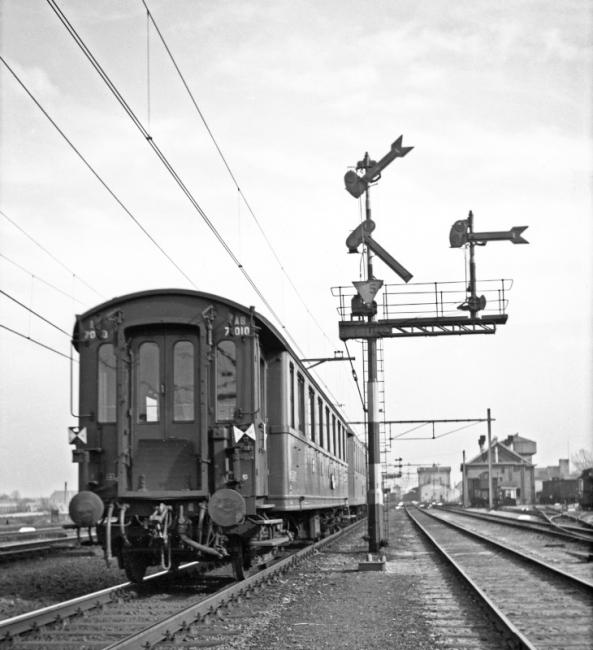Het inrijsein van station Gouda vanuit de richting Moordrecht met een passerende NS reizigerstrein met achteraan personenrijtuig AB 7010, een D-treinrijtuig, dus vermoedelijk betreft het een internationale trein uit Den Haag SS of Hoek van Holland.Gelet op de uitvoering van de seinarmen en de gebruikte schaarstellers dateert het sein uit de periode 1932-1937. De trein heeft de gebruikelijke sluitsein­borden; aan de aanwezigheid van de sluitseinen kon men zien dat de trein compleet was.