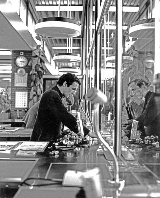 Een moderner plaatskaartenkantoor, ook te Amsterdam. De lokettist heeft aan zijn rechterhand de kasten waar de kaartjes klaarliggen; het apparaat dat omhoog steekt gebruikt hij om op de kaartjes van een datumstempel te voorzien. Daarnaast heeft hij nog diverse andere stempels tot zijn beschikking.