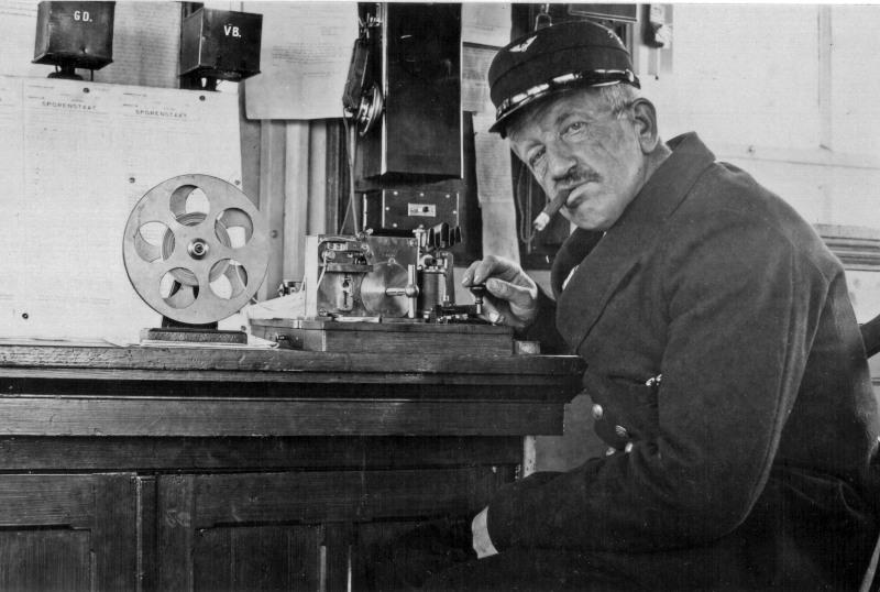 NS-medewerker zit aan het telegraaf-toestel; er werden berichten overgeseind in een code die bestond uit korte en lange piepjes, de zogenaamde Morse-code. De man heeft zijn hand aan de seinsleutel die hij moest indrukken om de signalen te verzenden. In het station waar het bericht via de draad naartoe werd gezonden, stond ook een dergelijk toestel. Aldaar werden de punten en strepen zichtbaar op de papierstrook zoals die links te zien is.Op de kasten linksboven is te zien: GD voor Gouda, VB voor Voorburg. Deze wachtpost kon met beide contact maken.