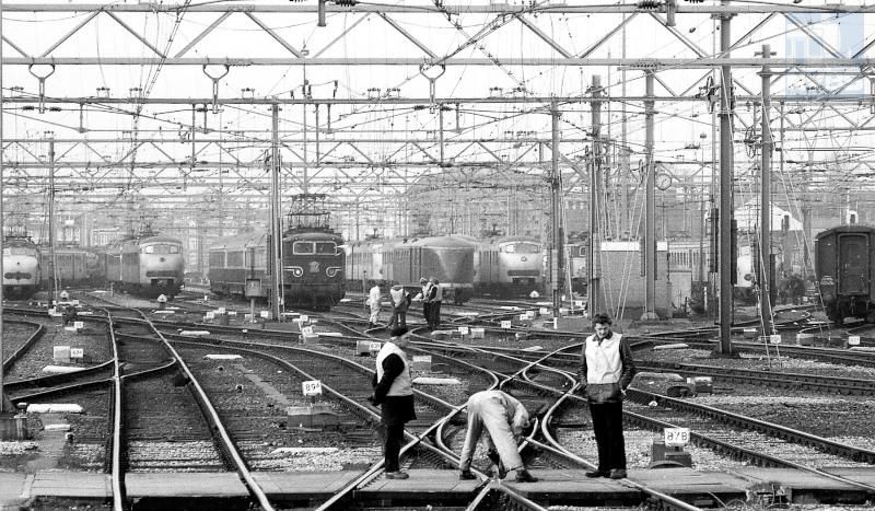 Op het Westelijk Eiland Amsterdam CS wordt aan een half Engels wissel gewerkt. Van links naar rechts: eentreinstel mat ′54 (Hondekop), een treinstel mat ′64 (Plan T of V), een nog blauwe eloc serie 1100 in oorspronkelijke uitvoering met een TEE naar Duitsland, een postrijtuig voor treinstellen een zogenaamde "Pec" (Postrijtuig voor elektrische tractie met wc), nog een treinstel mat′64 en een eloc serie 1100 in geel/grijs met botsneus.De foto is gemaakt door Frans Boom op 14 februari 1977.