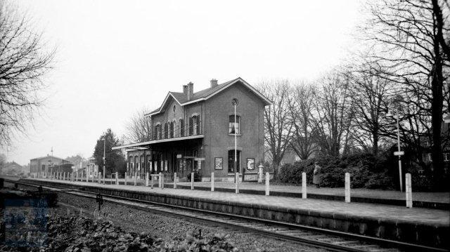 Gezicht op station Delden met op de achtergrond de goederenloods. Het station is een klassiek voorbeeld van een 4e klasse Waterstaatstation, ontworpen door architect K.H. van Brederode en in 1863 geopend. In 1904 werd er nog een vleugel aan het gebouw toegevoegd. Omdat het station nog altijd vrijwel in de oorspronkelijke staat verkeerde, behoorde het al in 1957 tot de collectie van bijzondere stationsgebouwen in Nederland. 24 december 1957. (Foto: J.A. Bonthuis - verz. NVBS Railverzamelingen - NEG530-015)