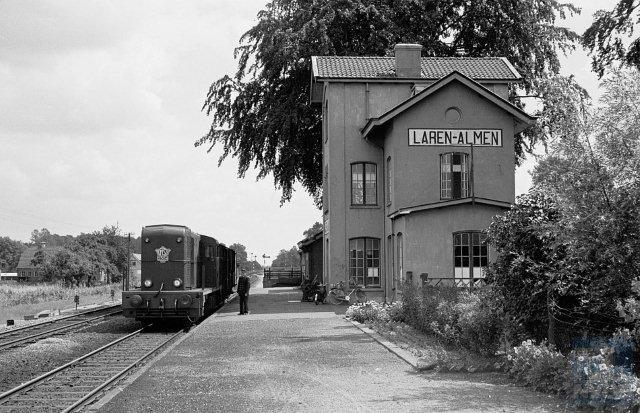 Station Laren-Almen startte zijn bestaan onder de naam station Laren. In 1925 werd het Laren-Almen. Het heeft niet mogen baten, in 1938 werd het station al gesloten. Een Waterstaatstation 5e klasse. Het oorspronkelijke gebouw bestond uit een twee verdiepingen hoog middengebouw met puntgevel en twee smalle even hoge vleugels. In 1901 werd een lage zijvleugel bijgebouwd en in 1914 het hoofdgebouw met een verdieping verhoogd. Foto: gezicht op de sporen en het net geschilderde stationsgebouw van Laren-Almen met rijwieltrein 4465 (Lochem - Zutphen) bestaande uit locomotief NS 2504 en twee gesloten goederenwagens op spoor 1 bij km 10,15. De blikrichting is oost (richting Hengelo). 24 juli 1960. (Foto: R. Ankersmit)