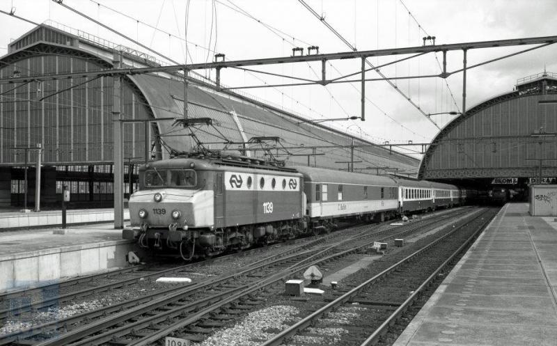 De internationale reizigerstrein getrokken door eloc 1139 staat even buiten de immense kap van het station Amsterdam CS klaar om naar het Zuidfranse Port Bou te vertrekken. Vergeleken met de vorige foto van dit type hebben de 1100-en hier een botsneus gekregen en een andere kleurstelling. Ook deze foto is van de hand van Bonthuis. 4 augustus 1989 (NVBS-fotonummer 539.600).